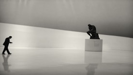 A person walks across a vast, empty, white space with a minimalist design. A large sculpture of a contemplative figure is positioned on a plinth on the right side of the image. The room is characterized by soft lighting and a sleek, modern aesthetic.