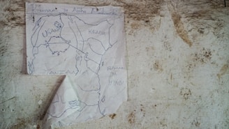 A hand-drawn map is taped to a textured, worn wall. The map displays parts of East Africa, with country names like Uganda, Kenya, and Tanzania clearly written. Below these, the Indian Ocean is labeled in Swahili as 'Bahari ya Hindi'. The paper is slightly crumpled and appears to be affixed to the wall with a single piece of tape.