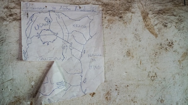 A hand-drawn map is taped to a textured, worn wall. The map displays parts of East Africa, with country names like Uganda, Kenya, and Tanzania clearly written. Below these, the Indian Ocean is labeled in Swahili as 'Bahari ya Hindi'. The paper is slightly crumpled and appears to be affixed to the wall with a single piece of tape.
