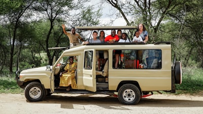 A group of people are gathered on and around a safari vehicle in a lush, wooded area. The vehicle is a beige off-road jeep with an open roof. Some individuals are seated inside while others are standing on the vehicle, with one person waving. The scene is lively and cheerful, suggesting a travel or adventure experience.