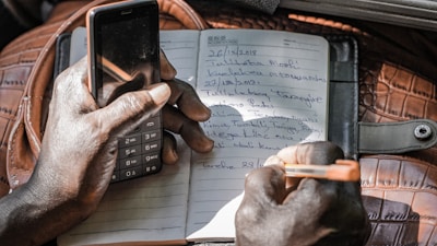 A person is holding a basic mobile phone in one hand while writing notes with an orange pen on a lined notebook. The notebook is placed on a brown textured bag or cover, with handwritten text visible on the pages.