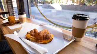 A croissant sandwich filled with cheese is placed on a tray lined with parchment paper on a wooden table. Alongside is a paper cup with a cardboard sleeve containing a hot beverage. In the background, another table holds a muffin and a similar cup of coffee. The scene is set beside a window, revealing a blurred view of a street outside.