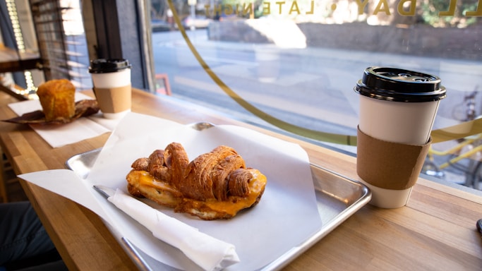 A croissant sandwich filled with cheese is placed on a tray lined with parchment paper on a wooden table. Alongside is a paper cup with a cardboard sleeve containing a hot beverage. In the background, another table holds a muffin and a similar cup of coffee. The scene is set beside a window, revealing a blurred view of a street outside.