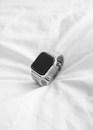 A sleek, modern smartwatch with a black screen and silver metallic band rests on an evenly wrinkled white fabric surface.