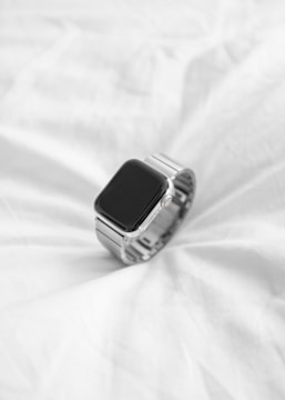 A sleek, modern smartwatch with a black screen and silver metallic band rests on an evenly wrinkled white fabric surface.