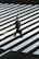 A person in a suit carrying a briefcase crosses a wide zebra pedestrian crossing. The geometric pattern of the white stripes contrasts sharply with the dark pavement.