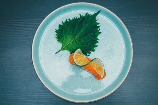 A minimalist food presentation featuring a piece of fresh salmon topped with two thin slices of lime, placed on a large green leaf. The elements are arranged on a round, light blue ceramic plate against a dark wood background.