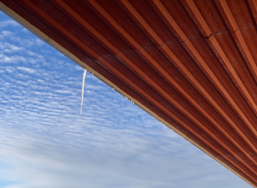 A corrugated metal roof extends diagonally across the image, contrasting with the blue sky. An icicle hangs from the edge of the roof, highlighting the cold weather. Puffy white clouds spread across the sky, adding texture and depth to the scene.