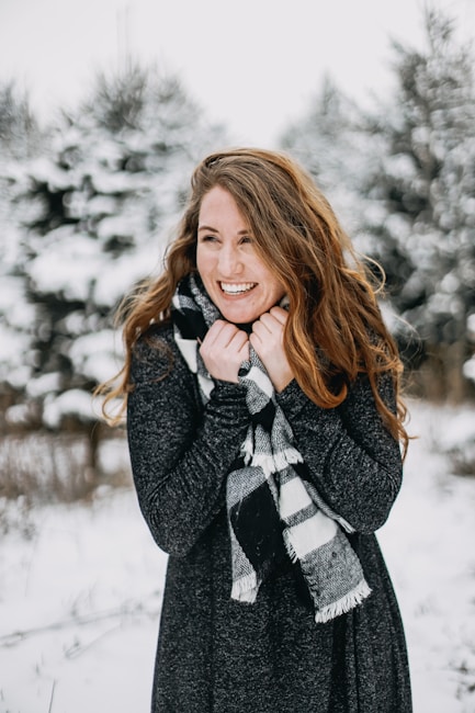 A person with long hair is standing outdoors in a snowy landscape, wrapped in a cozy black and white checkered scarf and wearing a dark sweater. Their expression is joyful and they appear to be enjoying the cold weather.