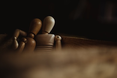 Two wooden mannequin figures sit closely together in a dimly lit and blurred foreground environment, suggesting a sense of companionship or comfort. The focus is on the mannequins, with one figure leaning gently towards the other.