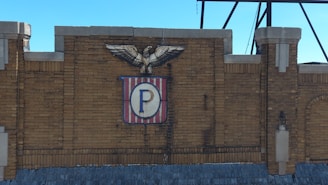 A brick wall with an emblem featuring an eagle perched on top. The emblem includes a shield with vertical red and white stripes and a central blue 'P'.