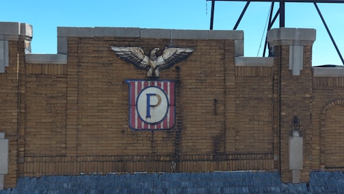 A brick wall with an emblem featuring an eagle perched on top. The emblem includes a shield with vertical red and white stripes and a central blue 'P'.