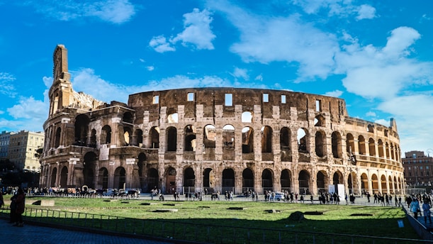 A large ancient amphitheater, the Colosseum, stands prominently under a vibrant blue sky scattered with clouds. The structure's arches and architectural details are distinct, showcasing weathered stone and a series of arches. People walk around the grassy area in front, highlighting the monument's grand scale.