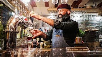 A bearded man wearing a beret and denim apron is pouring a beverage from a tap behind a bar. The background shows shelves with various bottles and neatly arranged hanging glasses.