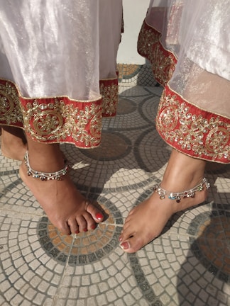 Pair of layered anklets featuring intricate patterns on a model’s bare feet near a floral arrangement