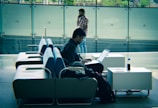 A person is sitting on a row of modern sofas using a laptop. The setting appears to be in a spacious, well-lit indoor area with a glass wall in the background. Another person is walking while talking on the phone in the background. A bottle and some papers are on a side table next to the seated individual.