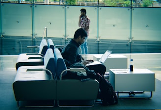 A person is sitting on a row of modern sofas using a laptop. The setting appears to be in a spacious, well-lit indoor area with a glass wall in the background. Another person is walking while talking on the phone in the background. A bottle and some papers are on a side table next to the seated individual.