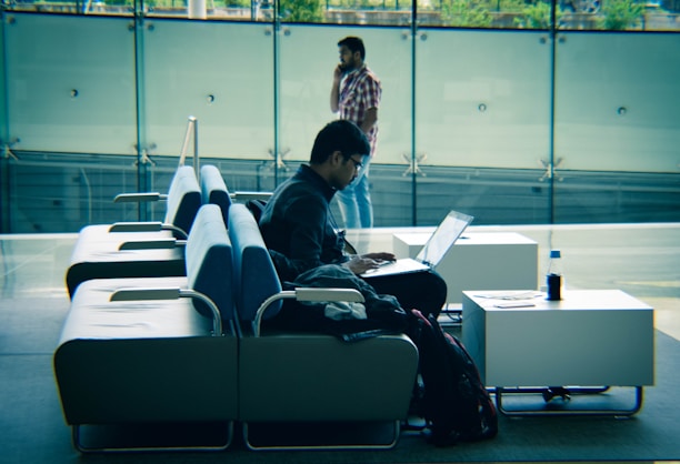 A person is sitting on a row of modern sofas using a laptop. The setting appears to be in a spacious, well-lit indoor area with a glass wall in the background. Another person is walking while talking on the phone in the background. A bottle and some papers are on a side table next to the seated individual.