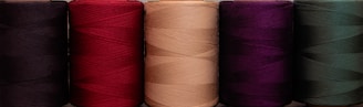 A row of five large spools of thread, each a different color, including black, red, beige, purple, and dark green. The threads are neatly wound, displaying a rich texture and vivid hues.