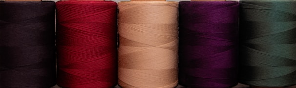 A row of five large spools of thread, each a different color, including black, red, beige, purple, and dark green. The threads are neatly wound, displaying a rich texture and vivid hues.