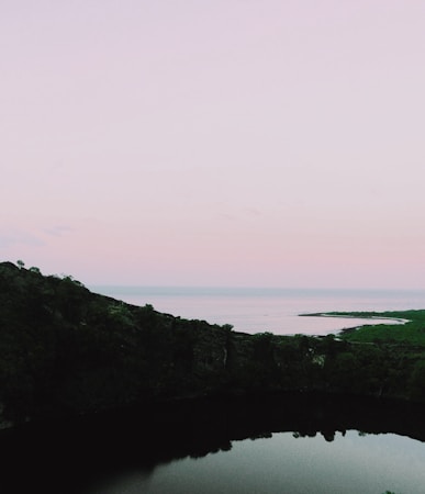 A serene landscape featuring a calm body of water surrounded by a lush green hillside. The sky above is a soft gradient of pastel colors, transitioning from light pink to pale blue, suggesting a sunset or sunrise. The distant horizon blends with the expansive water view, creating a peaceful and tranquil scene.