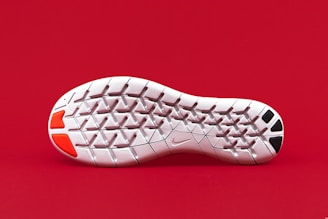 A close-up view of the sole of a white shoe with distinct geometric patterns. The sole has red and black patches on its heel area and a small logo in the middle. The background is solid red, making the sole stand out prominently.