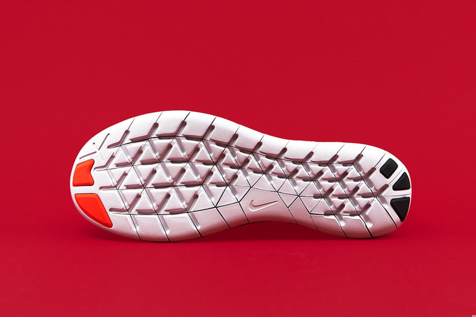 A close-up view of the sole of a white shoe with distinct geometric patterns. The sole has red and black patches on its heel area and a small logo in the middle. The background is solid red, making the sole stand out prominently.
