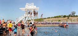 People enjoy a sunny day at a bustling outdoor swimming area with a large diving platform. Several individuals are diving into the water, while others are swimming or observing from the deck. A variety of people, including children and adults, are engaged in leisure activities. The backdrop includes trees, a grassy landscape, and a clear blue sky.