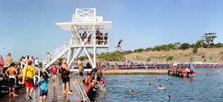 People enjoy a sunny day at a bustling outdoor swimming area with a large diving platform. Several individuals are diving into the water, while others are swimming or observing from the deck. A variety of people, including children and adults, are engaged in leisure activities. The backdrop includes trees, a grassy landscape, and a clear blue sky.
