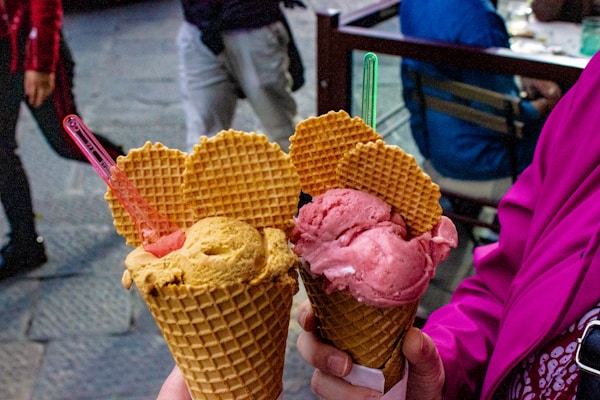 Two waffle cones are filled with ice cream topped with heart-shaped waffle pieces and colorful plastic spoons. One cone seems to have a scoop of yellow and pink ice cream, while the other features a pink scoop. The background shows people walking by, creating a casual outdoor setting.