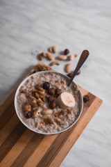 A bowl of oatmeal topped with various nuts and a slice of banana sits on a wooden board. A spoon is placed in the bowl, and some scattered nuts can be seen in the background on a light-colored surface.