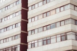 A multi-story residential building with rows of rectangular windows framed in red. The facade is predominantly light gray with concrete textures, and there are visible reflections on some of the windows.