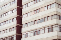 A multi-story residential building with rows of rectangular windows framed in red. The facade is predominantly light gray with concrete textures, and there are visible reflections on some of the windows.