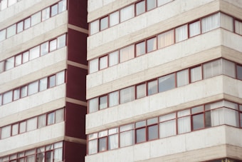 A multi-story residential building with rows of rectangular windows framed in red. The facade is predominantly light gray with concrete textures, and there are visible reflections on some of the windows.