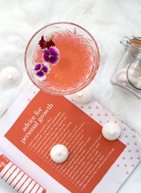 A delicate cocktail in an intricately designed glass is garnished with vibrant purple and pink flowers, set atop a white surface. Nearby, there is an open magazine displaying an orange page with the title 'advice for personal growth'. Surrounding the magazine and glass are small, white meringue-like treats. On the right, a glass jar with a latched lid contains more of these treats.