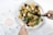 A plate filled with a fresh salad including leafy greens, cubes of sweet potato, feta cheese, and walnuts. A person is using a knife and fork to cut into the salad. An open magazine with business-related content can be seen beside the plate, placed on a white surface.