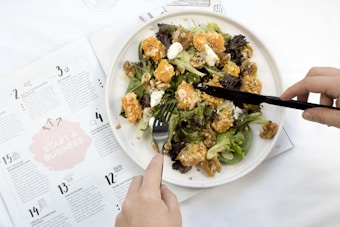 A plate filled with a fresh salad including leafy greens, cubes of sweet potato, feta cheese, and walnuts. A person is using a knife and fork to cut into the salad. An open magazine with business-related content can be seen beside the plate, placed on a white surface.