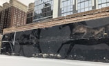 A large mural depicting two black horses in motion is painted on an old brick wall. The urban backdrop consists of modern high-rise buildings with reflective glass windows and older, more traditional brick buildings. The ground is covered with a thin layer of snow.