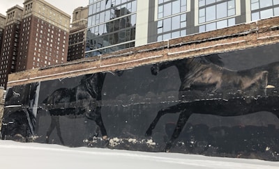 A large mural depicting two black horses in motion is painted on an old brick wall. The urban backdrop consists of modern high-rise buildings with reflective glass windows and older, more traditional brick buildings. The ground is covered with a thin layer of snow.