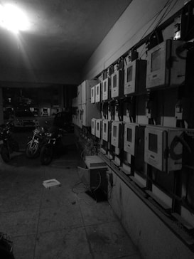 A dimly lit room contains a series of electric meters mounted in a row on a wall. Several motorcycles are parked alongside the wall in a somewhat cluttered space. A single light bulb provides illumination from the ceiling, casting shadows around the meters and vehicles. Cables are seen hanging from the meters, and a box lies on the ground, contributing to the disarray.