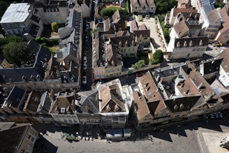 An aerial view of a densely packed residential area featuring traditional European architecture. The buildings have steep, tiled roofs and pastel-colored walls. Narrow streets and alleys separate the buildings, with some greenery visible. Outdoor seating arrangement and a few individuals can be seen at street level.