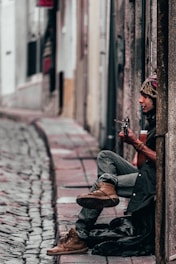 A person sits against a wall on a cobblestone street, playing a guitar. The individual is dressed in casual attire, including a beanie and boots. The street is narrow and lined with buildings, creating an urban atmosphere.