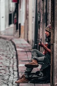 A person sits against a wall on a cobblestone street, playing a guitar. The individual is dressed in casual attire, including a beanie and boots. The street is narrow and lined with buildings, creating an urban atmosphere.