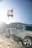 A person is captured in mid-air performing a jump near a rugged mountain terrain. Below the person, a silver SUV is parked on a rocky surface, suggesting an adventurous setting. The sunlight adds a dramatic flair, highlighting the action taking place.