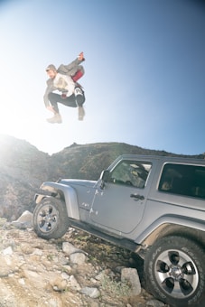 A person is captured in mid-air performing a jump near a rugged mountain terrain. Below the person, a silver SUV is parked on a rocky surface, suggesting an adventurous setting. The sunlight adds a dramatic flair, highlighting the action taking place.