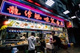 A bustling street food stall is illuminated by vibrant neon lights. The neon sign is written in an East Asian language, and a variety of food items and prices are displayed on an overhead menu. Multiple people are standing in front of the stall, some engaged in purchasing food. The stall itself is packed with food items and cooking utensils, creating a lively and dynamic atmosphere.