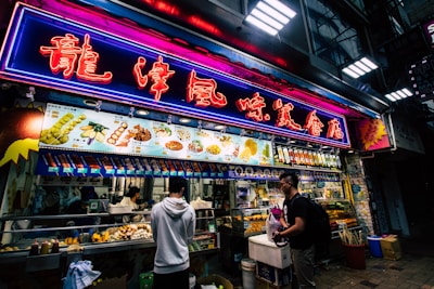 A bustling street food stall is illuminated by vibrant neon lights. The neon sign is written in an East Asian language, and a variety of food items and prices are displayed on an overhead menu. Multiple people are standing in front of the stall, some engaged in purchasing food. The stall itself is packed with food items and cooking utensils, creating a lively and dynamic atmosphere.