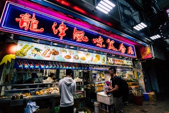 A bustling street food stall is illuminated by vibrant neon lights. The neon sign is written in an East Asian language, and a variety of food items and prices are displayed on an overhead menu. Multiple people are standing in front of the stall, some engaged in purchasing food. The stall itself is packed with food items and cooking utensils, creating a lively and dynamic atmosphere.