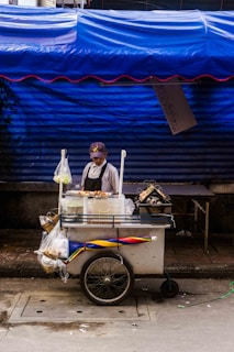 A street vendor stands behind a small food cart, which is equipped with a display of skewered food items. The vendor wears an apron and a cap. The cart is adorned with a colorful striped umbrella and several plastic bags containing various goods. This scene is set against a backdrop of a bright blue tarp, providing a vivid contrast to the street setting.