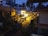 A residential street is dimly lit with warm yellow lights, surrounded by houses and trees. The buildings have balconies and are closely situated. Several palm trees are visible in the background under an evening sky.